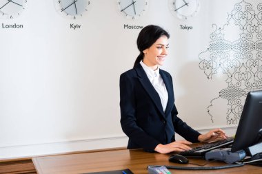 happy brunette receptionist in formal wear standing near computer monitor in hotel  clipart