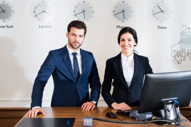 cheerful woman near serious receptionist in suit standing at reception desk  clipart