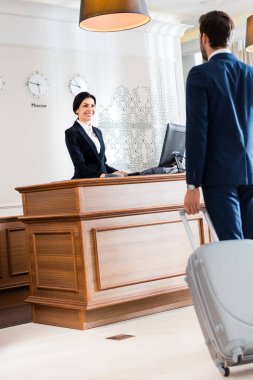 selective focus of attractive receptionist looking at businessman walking with luggage  clipart