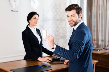 selective focus of cheerful businessman holding credit card near serious receptionist  clipart