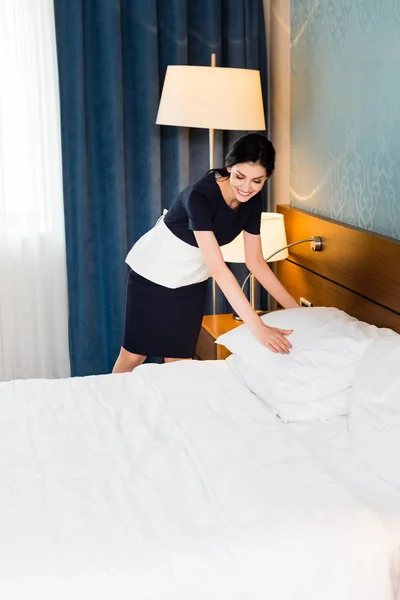 ホテルの部屋でベッドの上に白い枕に触れる幸せな家政婦 — ストック写真