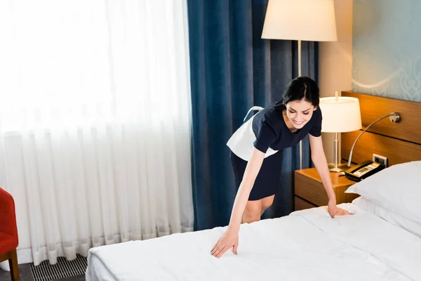 happy housemaid touching white bedding in hotel room
