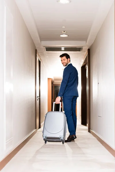 ホテルの廊下でスーツケースを持って立っている間に笑顔の陽気なあごひげの男 — ストック写真
