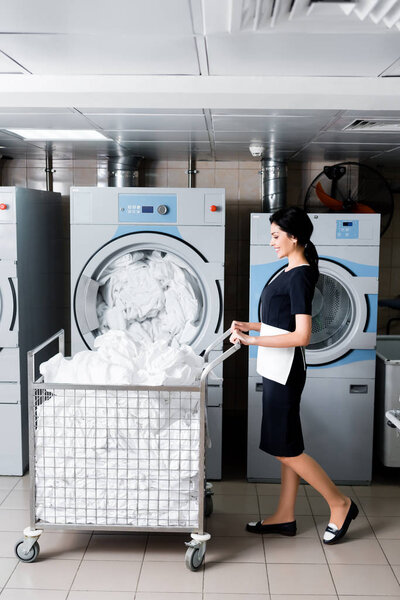 счастливая горничная смотрит на грязное постельное белье, стоя возле металлической тележки
 