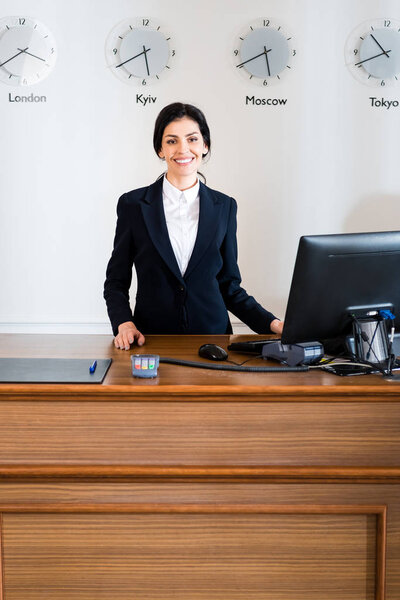 веселая брюнетка секретарша в формальной одежде стоит рядом с монитором компьютера в отеле
 