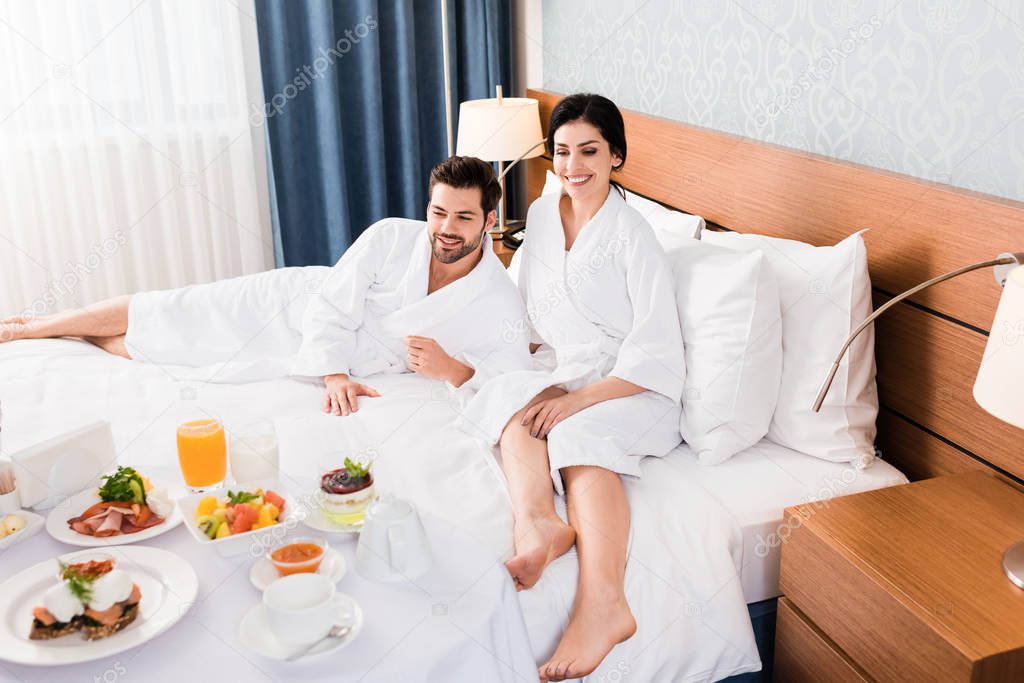 happy man and woman with barefoot looking at breakfast in hotel room 