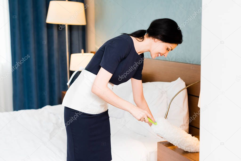 happy maid holding duster and cleaning hotel room 