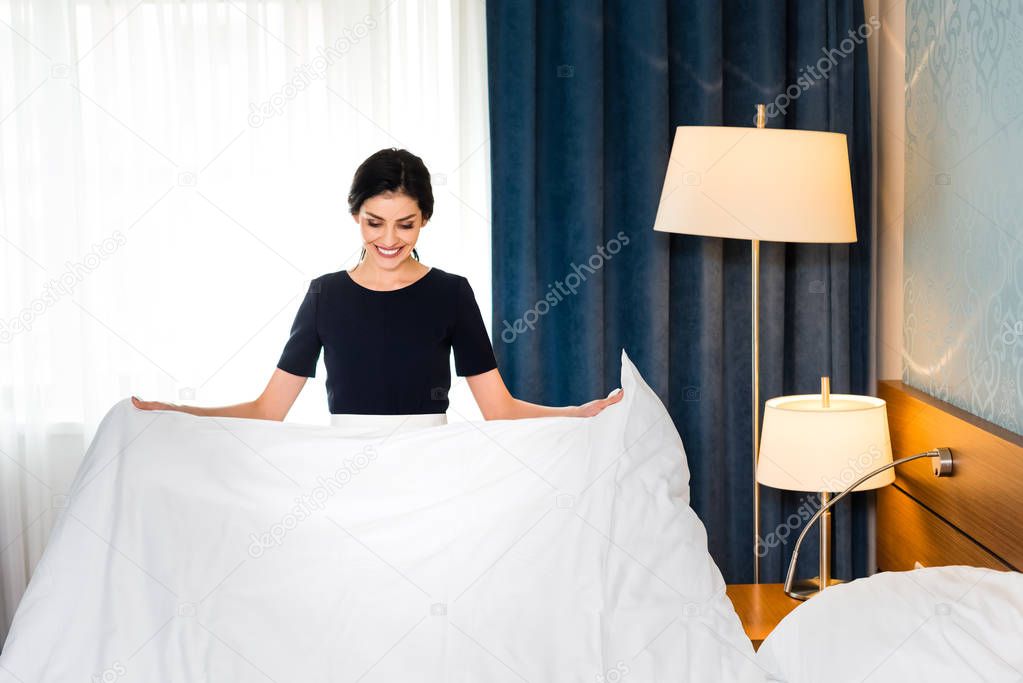 cheerful housemaid holding white bed sheet in hotel room 