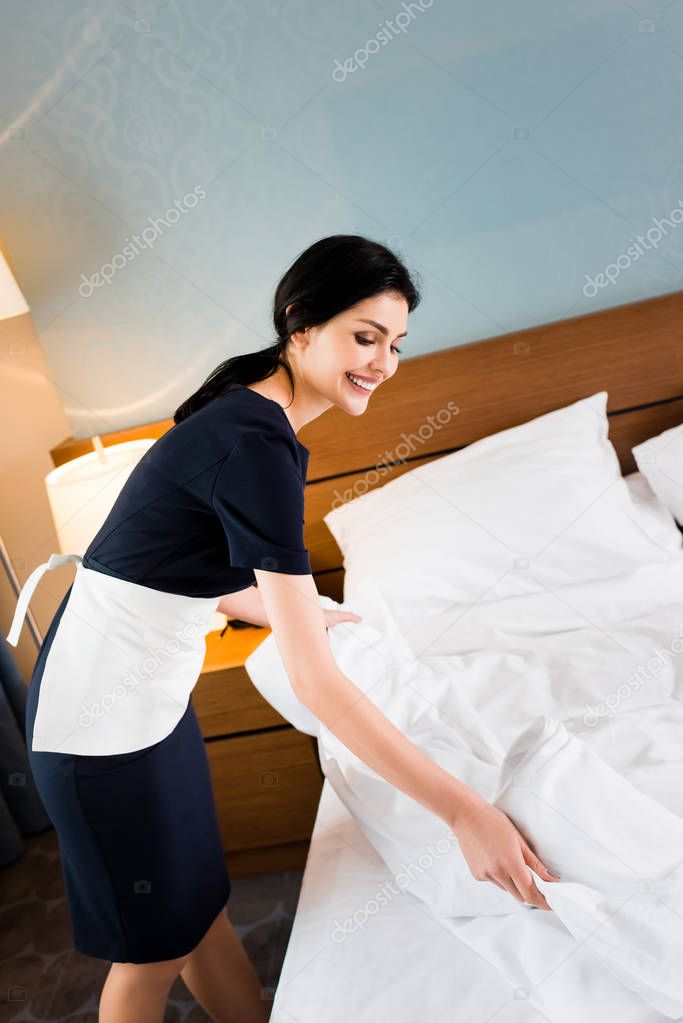 happy brunette housemaid touching white bedding in hotel room 