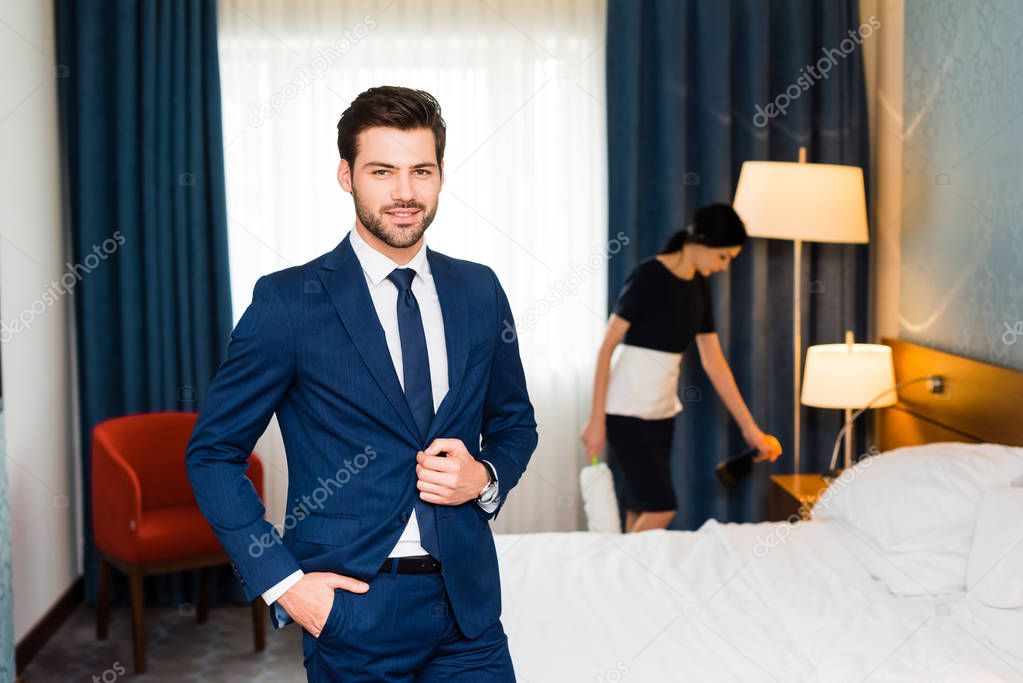 happy receptionist standing with hand in pocket near maid in hotel room 