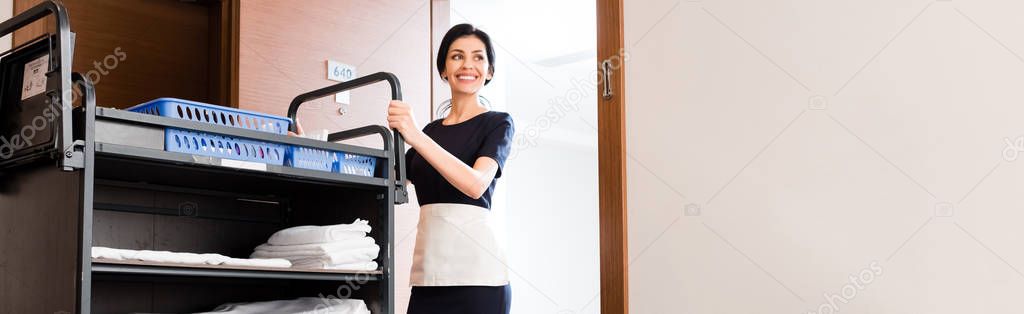 panoramic shot of cheerful housemaid in uniform standing near cleaning trolley with white towels  