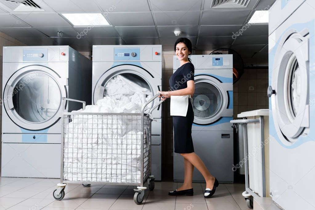 cheerful maid standing near metallic cart with bedding in laundry 