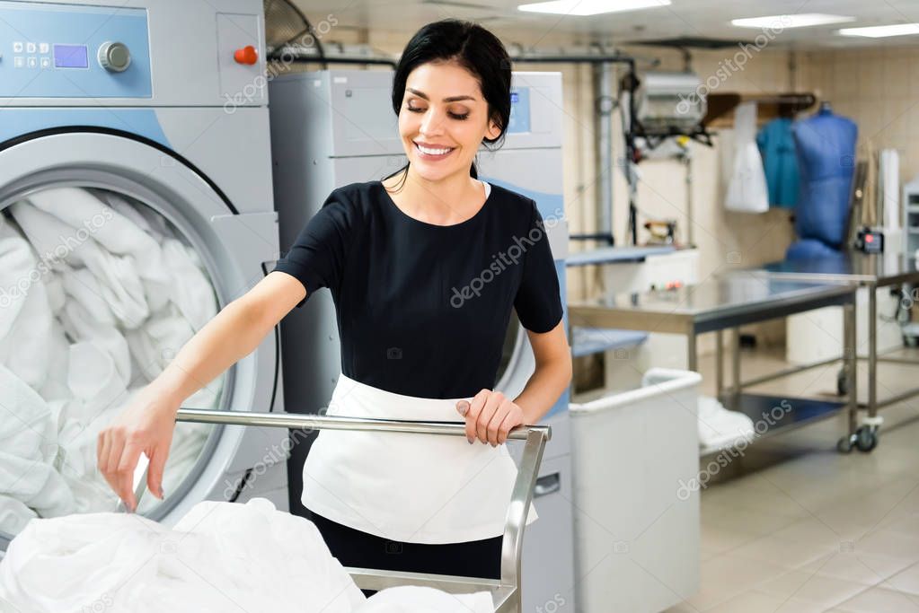 selective focus of smiling housemaid near basket with white bed sheets