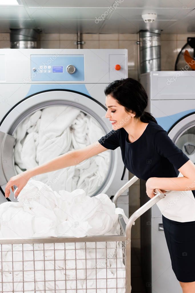 cheerful maid touching bed sheets while standing near washing machines in laundry 