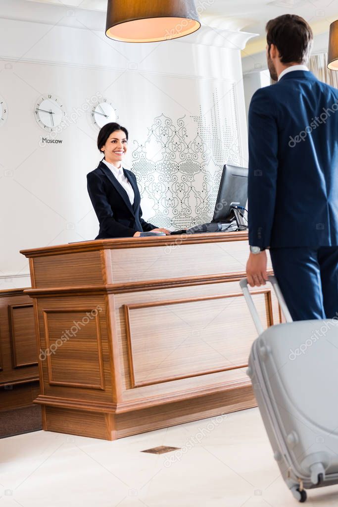 selective focus of attractive receptionist looking at businessman walking with luggage 