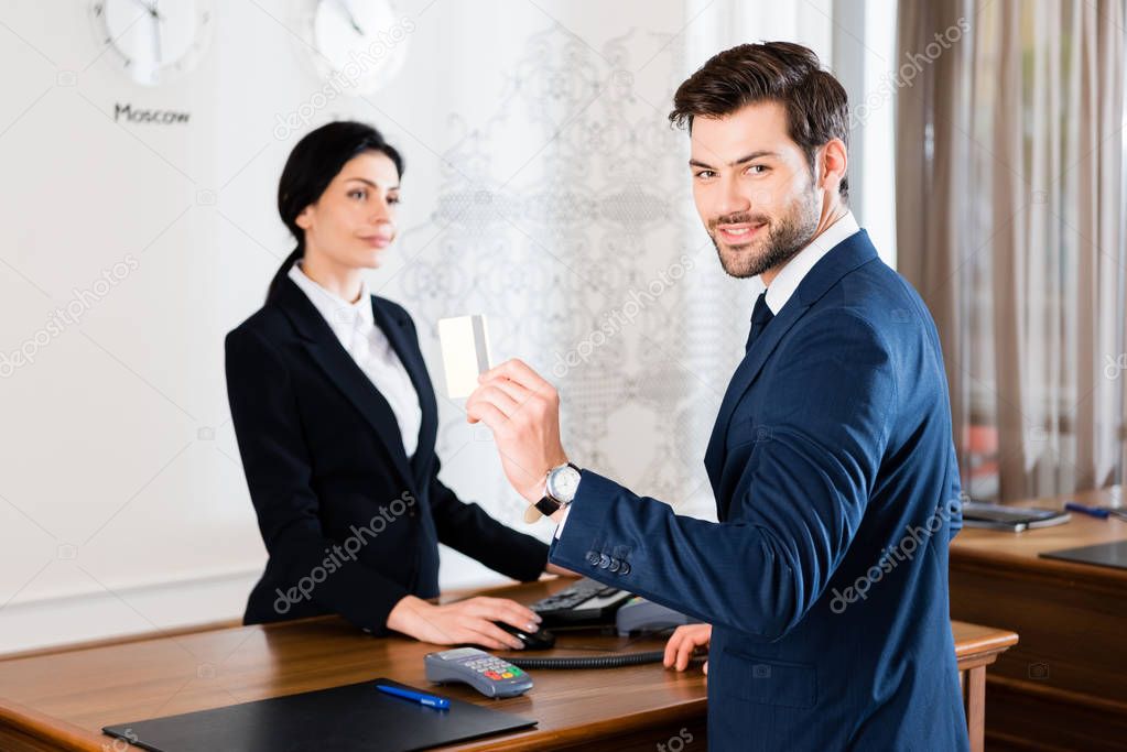 selective focus of cheerful businessman holding credit card near serious receptionist 