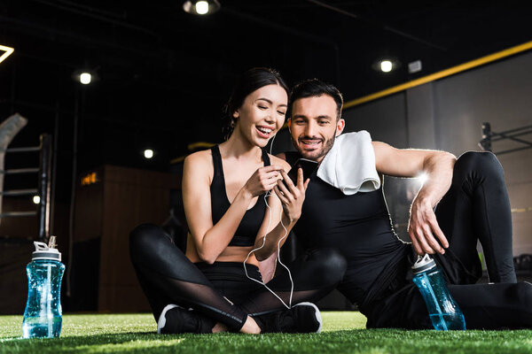 низкий угол зрения веселой женщины в спортивной одежде с помощью смартфона рядом мужчина сидит на траве
 