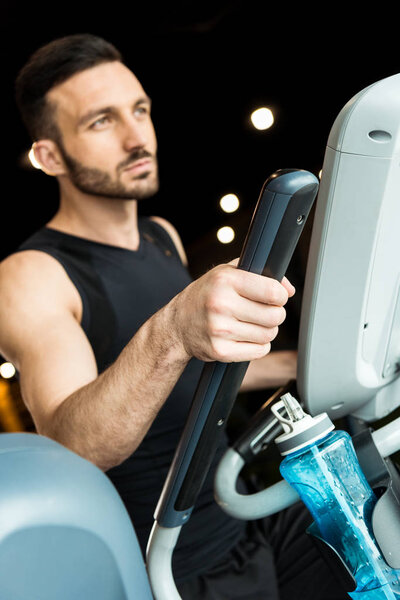 selective focus of sport bottle near athletic man working out on exercise bike 