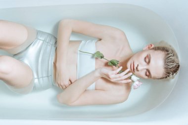 top view of tender woman with closed eyes gently holding flower in transparent water in bathtub clipart