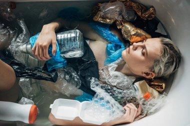 top view of young sad woman lying in bathtub with trash, environmental pollution concept clipart
