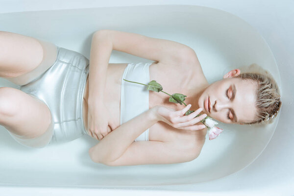 вид сверху на нежную женщину с закрытыми глазами, нежно держащую цветок в прозрачной воде в ванной
