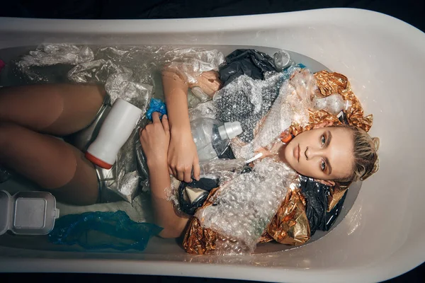 年轻女子躺在浴缸与垃圾隔离在黑色 环境污染概念的顶视图 — 图库照片