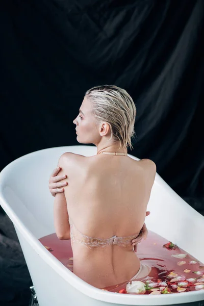 背面的金发性感湿女人坐在浴缸与粉红色的水和玫瑰花瓣 — 图库照片