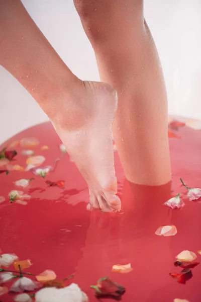 赤脚妇女站在白色浴缸与粉红色的水和花瓣的裁剪视图 — 图库照片