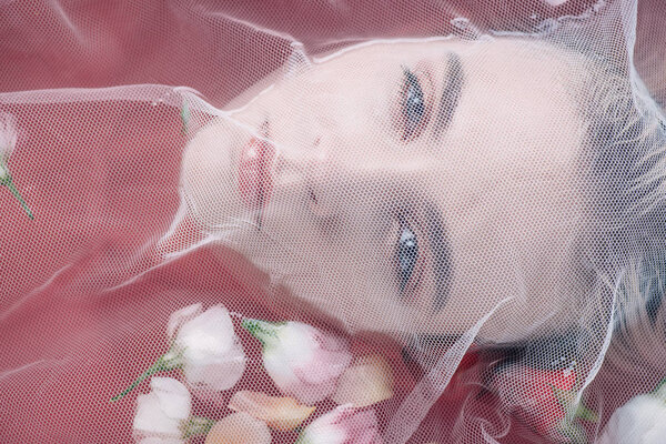 портрет красивой женщины, покрытой белой сеткой ткани в розовой воде с лепестками роз
 