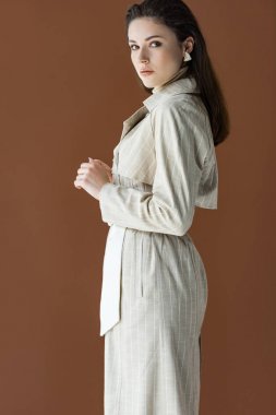 side view of trendy woman in stylish trench coat isolated on brown, looking at camera clipart