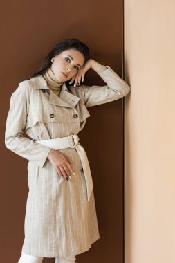 stylish woman in trendy trench coat standing on brown background, looking at camera clipart