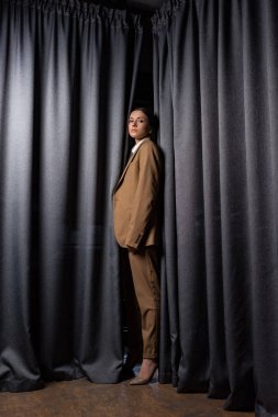 side view of fashionable model in suit standing on dark grey curtain background, looking at camera clipart