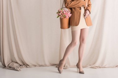 cropped view of model in trendy wear carries flowers on shoulder bag, standing on curtain background clipart