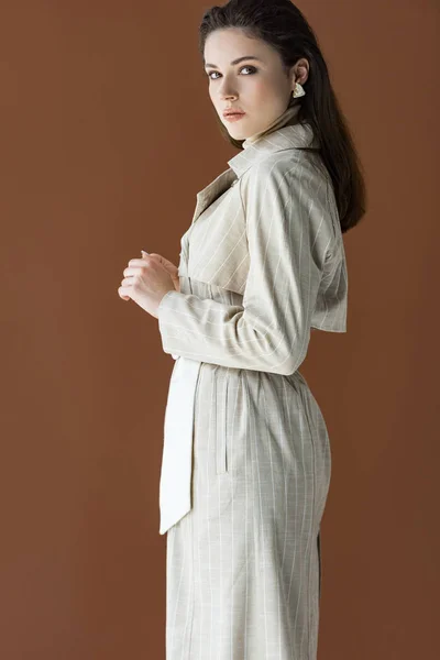 side view of trendy woman in stylish trench coat isolated on brown, looking at camera