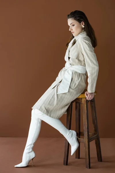 茶色に隔離された椅子に座っているトレンチコートのファッショナブルな女性の側面図 — ストック写真