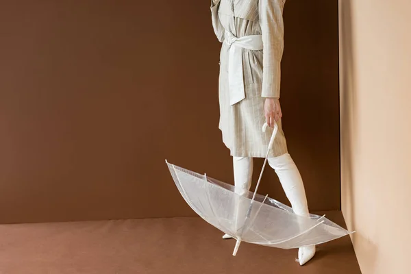 茶色の背景に立つ傘を持つベージュのトレンチでモデルのトリミングビュー — ストック写真