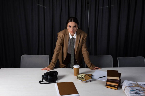 Взрослая деловая женщина, стоящая за столом в офисе, глядя в камеру
