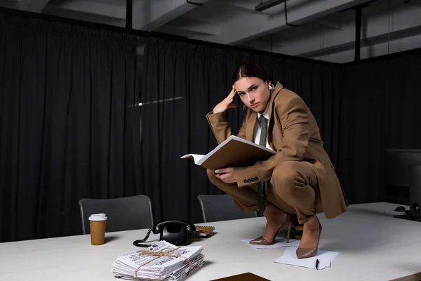 stylish businesswoman in suit sitting on table, holding documents and looking at camera