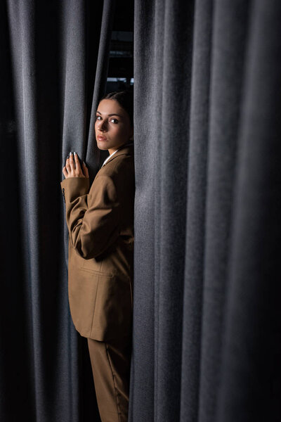 side view of successful woman in suit standing on dark grey curtain background, looking at camera