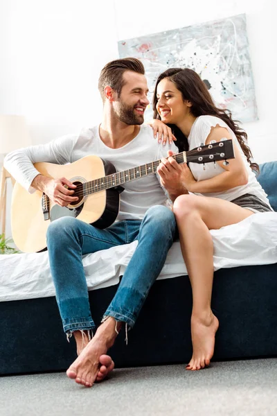침대에서 행복한 여자를 보면서 어쿠스틱 기타를 연주하는 — 스톡 사진