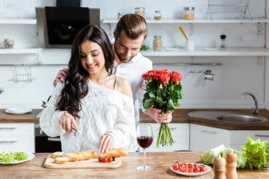 gülümseyen çift mutfakta birlikte ayakta kadın doğrama tahtası ve adam kırmızı gül tutan ekmek kesme