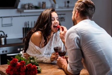 gül buketi ile ahşap masada kırmızı şarap bardakları ile çift, çilek ile erkek besleme kadın