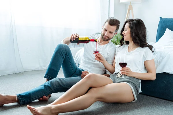 彼氏は寝室の床にガールフレンドと座って メガネのボトルから赤ワインを注ぐ — ストック写真