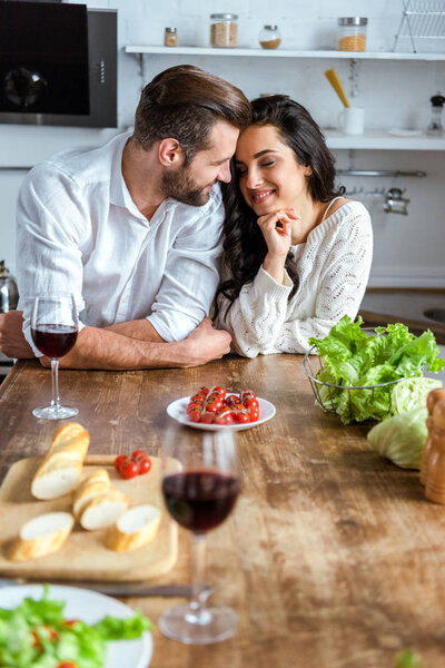 dreamy tender couple near wooden table with red wine, cherry tomatoes, bread and salad