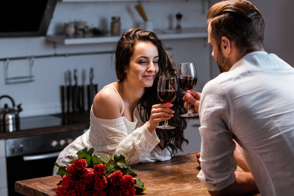 man and woman clinking with glasses of red wine at wooden table with bouquet of roses 