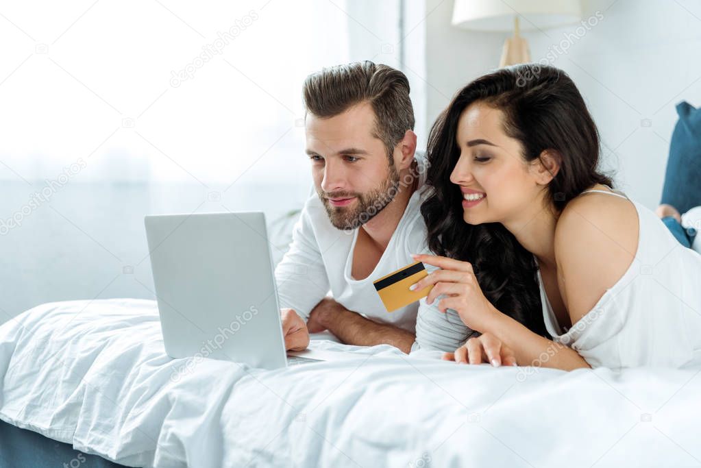 happy couple using laptop together and lying in bed while woman holding credit card, illustrative editorial