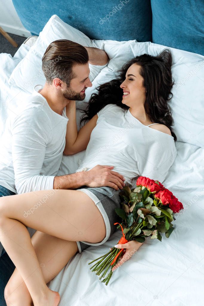 overhead view of happy couple lying in bed with bouquet of red roses