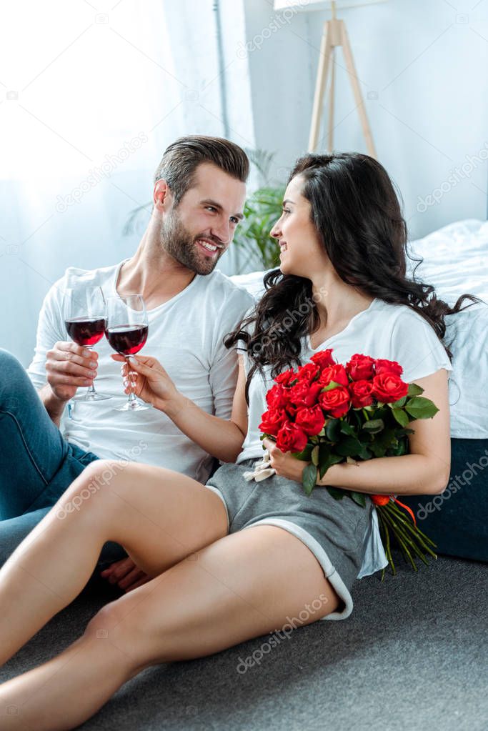 couple clinking with glasses of red wine and woman holding red roses and looking at man in bedroom