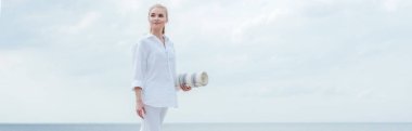 panoramic shot of happy young blonde woman standing near sea and holding yoga mat  clipart