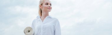 panoramic shot of cheerful young blonde woman standing near sea and holding yoga mat  clipart